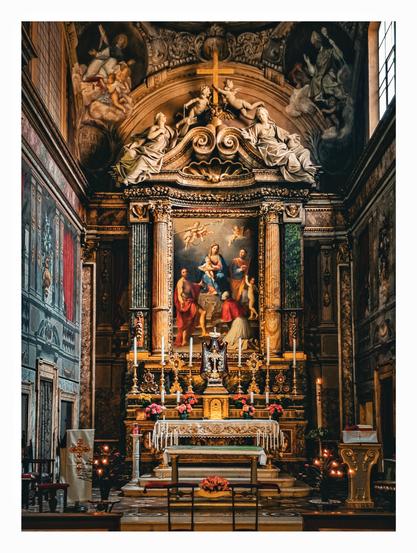 A colourful medieval church altar.  big painting of Mary holding Jesus surrounded by religious figures behind the altar table. Statues of angels adorn the top of the painting.