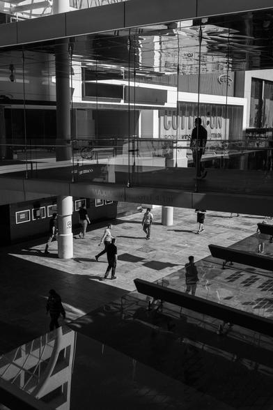 Black and white photo of Perth city with people walking on different levels. The scene includes reflections on glass surfaces and a visible sign in the background.