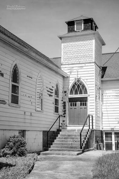 A church with double doors and a peaked stain glass window over it in a short bell tower in a corner of two parts of the building.  A sidewalk and stairs lead to the door.