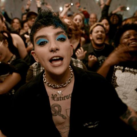 A white man with short, spiked black hair and an open black button-down shirt sings joyfully into the camera. He is wearing teal eye shadow and has tattoos across his chest. Behind him is a crowd of people singing joyfully.