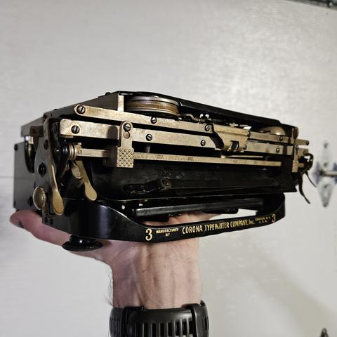 a corona typewriter, collapsed. it is black and brass colored, with gold lettering.