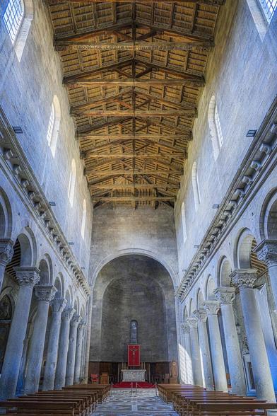 The narrow nave of the Viterbo Cathedral. A central aisle is flanked by rows of wooden pews. Rows of columns on either side of the nave have elaborate capitals. A simple altar in an alcove is at the far end. The ceiling is exposed wood.