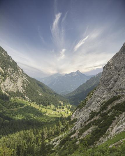 A sweeping alpine valley in Gesäuse National Park, Austria, with steep rocky slopes framing a lush green forest below. A scattering of traditional alpine buildings rests at the valley floor, surrounded by conifer trees. In the distance, rugged peaks rise through a soft haze, while delicate, wispy clouds fan out dramatically across a blue sky, adding a sense of motion and wonder to the tranquil mountain scene.