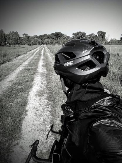 A bike rider seen from behind looking at a gravel road. The road is completly straight. We can see trees in the very background where leads the road. 
Picture is black and white