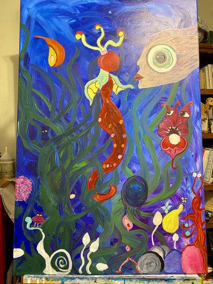 Oh my gosh, where to begin? It is a very large canvas with a background reminiscent of a kelp forest. There are many odd creatures, predominantly a mermaid with an apple for a head, a human headed fish with a univer spiraling into a black hole in her large oversized eye, a clam of sorts with tentacles and teeth and a whole host of undefined things which are still white spaces. It’s a very strange painting. It’s sitting on an easel, which is positioned in front of some book shelves.