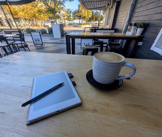 A striped ceramic mug filled with coffee sits on a saucer on a wooden table. Next to it is a digital note-taking device with a stylus placed on top. The setting is an outdoor cafe with tables and chairs in the background and a view of a street lined with trees.