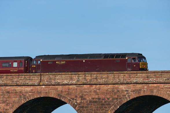 Close up shot of a maroon diesel with the logo "WEST COAST" on it mustard follows behind a maroon coach over a viaduct. In the background is blue sky with some cloud.