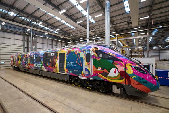 A brightly painted Avanti West Coast train is parked inside a spacious, well-lit maintenance depot. The train's exterior is covered in a vivid and dynamic mural featuring bold colors like pink, purple, green, and yellow, with stylized illustrations of people, musical notes, flowers, and abstract patterns. On the side near the front, the words "AVANTI WEST COAST" and the train number "807 010" are displayed within a blue panel. Near the rear of the train, the phrase "TOGETHER WE ROLL" is visible amidst the artwork. The train’s front end is signed by the artist, whose signature reads "Rinako Carbó" in flowing script. The depot has high ceilings, exposed beams, industrial lighting, and multiple tracks set into the concrete floor.