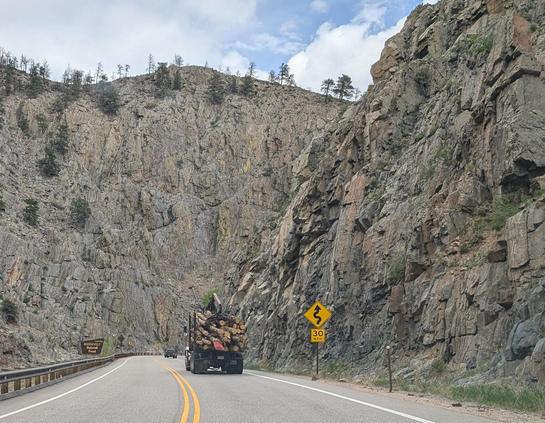 Road set in a deep gorge surrounded by the high foothills of the Rocky Mountains. Grey rock, a few trees, sign indicating 30 mph curve ahead. Truck with a load of fresh logs