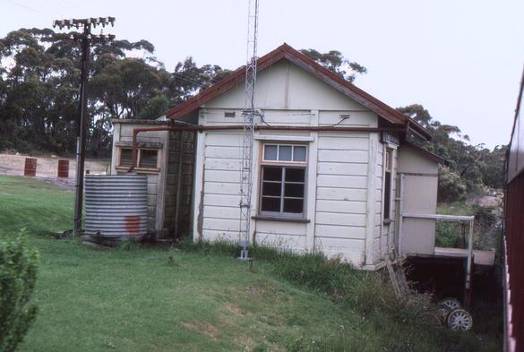 Snapped through an open window on the left hand side of a passenger train, we see a ground level signal box with peaked roof, corrugated iron water tank and fibro cement outbuilding to the left.  A staff exchange platform projects from the right towards the train which is just visible at far right.