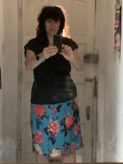 A middle aged woman wearing a black top and a turquoise A line knee length skirt with giant red and pink flowers on it with grey and black leaves