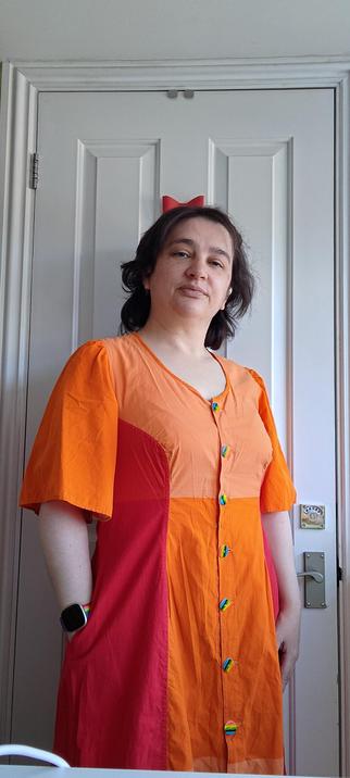 Middle aged white woman with dark hair standing in front of a white door wearing a colour block dress in shades of red and orange. It has rainbow buttons and she has a rainbow strap on her wristwatch.