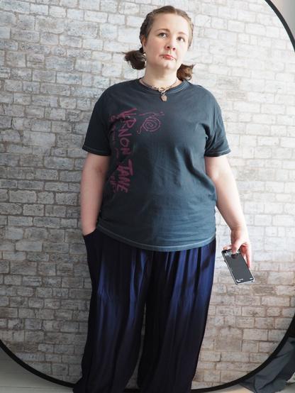 Photo of me, a cubby white woman-shaped type, with short brown hair in low bunches, wearing a 'unisex' cut t-shirt in a faded dark grey with blue undertones, and purple print 'Vernon Jane's 'Chestpains and Sidepieces' down one side. With baggy flowy dark blue (with purple undertones) trousers. And a short silver chain collar with a central o-ring from which dangles a silver coffin.