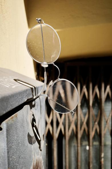 A close-up photo shows a pair of round, vintage-style eyeglasses mounted on a metal box, with one lens tilted upward and both lenses speckled with dirt, casting distinct shadows in bright sunlight.