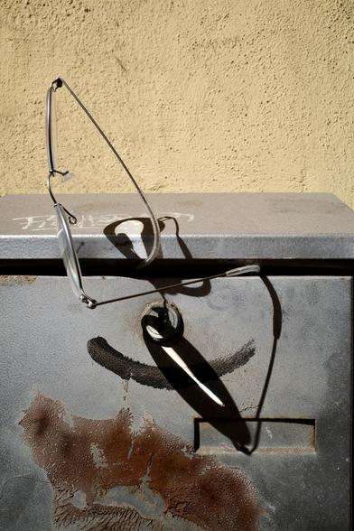 A pair of thin, metal-frame glasses lies atop a weathered, rust-streaked metal box against a textured yellow wall, casting a sharp, looping shadow in the sunlight.