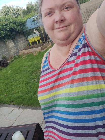 A fat white woman takes a selfie trying to show off her dress. The dress is sleeveless with a scoop neck. It's grey with rainbow stripes, you can almost see the matched patch pocket. She's in a garden with lawn and a greenhouse behind her
