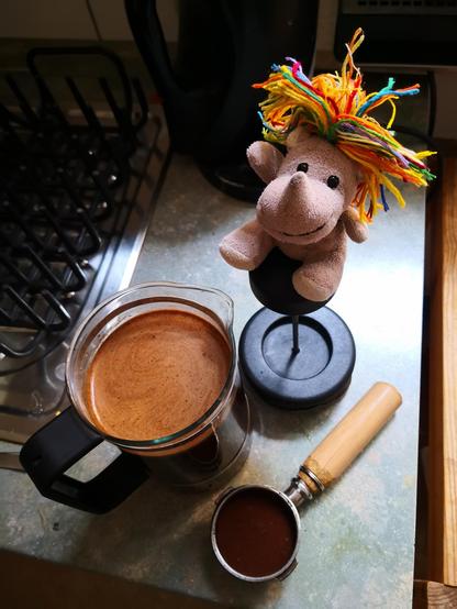 Das kleine Plüscheinhorn Carola sitzt auf dem Pressstab der French Press, daneben der aufgegossene Glasbehälter. Schon vorbereitet der mit frisch gemalenem und zusammengepressten Kaffee gefüllte Siebträger im Griff.