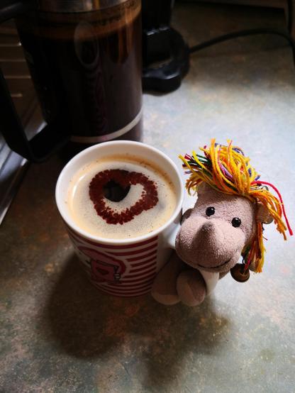 Das kleine Plüscheinhorn Carola hat sich in den Griff der Kaffeetasse geklemmt, auf der Crema ein Kakaoherz. 
Fertig zum Servieren!