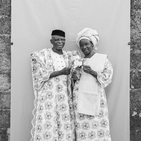 a black-and-white photograph in a square format of two Nigerians in traditional patterned garments, standing in front of a panel of fabric that has been installed on an exterior wall