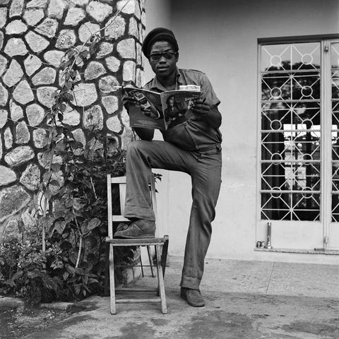 a black-and-white photograph in a square format of a Black man standing outside of a house, wearing fashionable 1970s apparel, standing with one leg up on the seat of a chair and holding a magazine