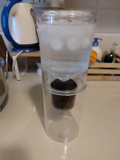 A photo of a vertical glass cold brew dripper, with frozen water and ice on top, dripping through some grounded coffee.