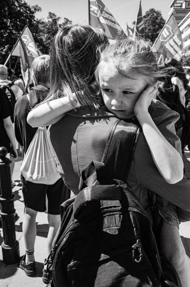 A young girl is carried through a crowd at a Labour Day march. She holds her hands over her ears and has a pained look on her face.