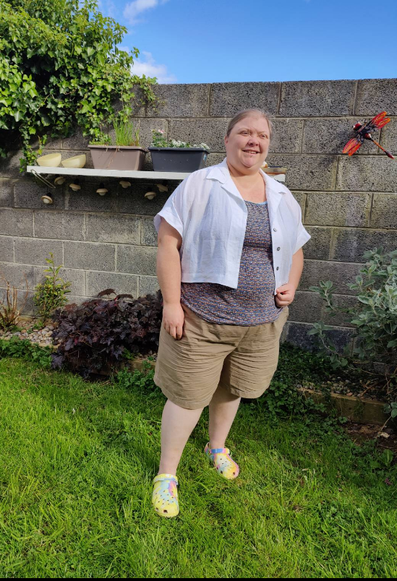 A fat white woman squints in the sun in a sunny garden. She is wearing a cropped white shirt with boxy short sleeves over a grey blue tank top. She's also wearing khaki coloured shorts and pastel rainbow crocs. There's a shelf on the wall with potted plants and a red metal dragonfly nailed to the wall. There's a raised bed with a dark heuchera among other plants. The top of the wall gives way to a bushy ivy and blue sky with a hint of cloud.