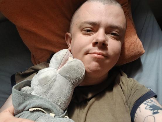 A white trans masc lying in bed, in a green t shirt and shaved head, hugging a stuffed hippo