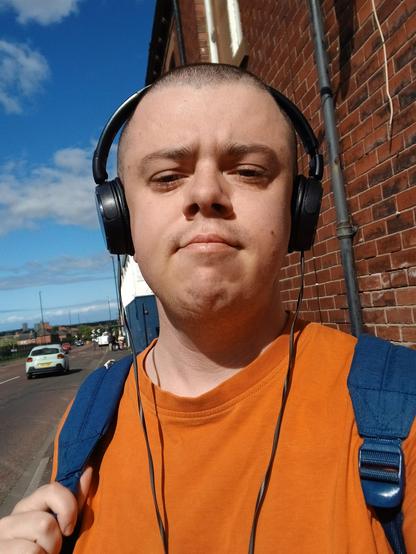A white transmasc in an orange t shirt, with black headphones, a blue rucksack, and a shaved head. He's stood in front of a brick wall