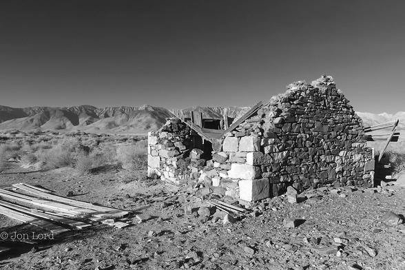 This Is A Black And White Landscape Photo In Landscape Format. Around Much Of The Image Is A Flat, Rough And Stoney Desert With Scattered Sparse Vegetation. To The Right Of Centre Is The Ruin Of A Long Abandoned Stone House. The Building Is Constructed From Stones And Rocks Of Various Sizes, The Larger And More Square Rocks Being In The Corners. The Roof Was Made From Wood And The Decaying Remains Are Scattered Inside The Structure. There Is A Pile Of Scattered Wood Planks To The Left. In The Distance Some Tens Of km Away Is A Tall Mountain Ridge Stretching Across The Image With A Clear And Cloudless Sky Above. 
The Location Is East Of Owens Lake In California. The Mountains: The Eastern Sierras (2016).