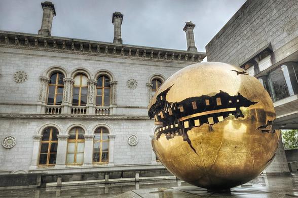 A large, golden, partially deconstructed sphere sculpture stands in front of a historic, light-colored stone building with arched windows on the Trinity College Dublin campus.