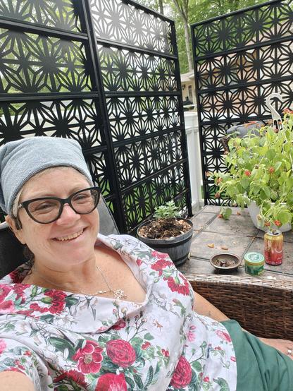 I am sitting outside, with a metal geometrical privacy screen behind me. I am wearing a shawl collared top, white with red carnations and green foliage and dark green shorts. Just ignore the "sexy" towel over my wet hair