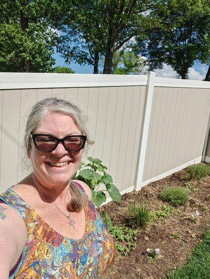Picture shows me from the boobs up, v neck, strappy dress in a fake rainbow marbleized print. Behind me is a row of plants
