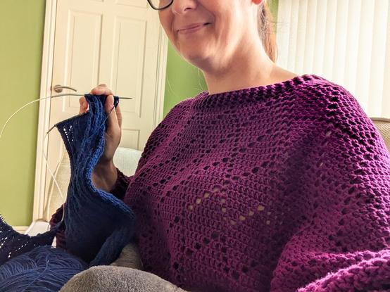 Cropped selfie of my general torso area. I'm sitting down and smiling slightly, wearing a long sleeved and aggressively purple crocheted jumper with an all-over diamond stitch pattern. I'm holding up a navy blue lacy cardigan knitting project, but no real detail is visible.