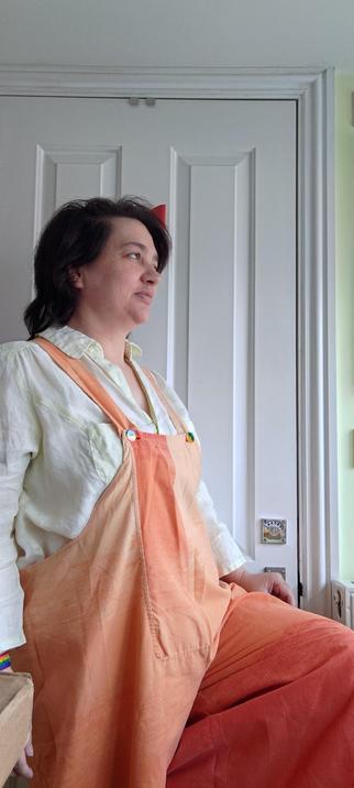 Me standing in front of a white door with one knee up to show the colour variation in these yellowy peachy orangey dungarees.