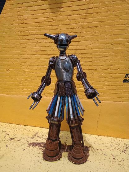 Statue of a robot made of scrap metal.  Yellow brick wall behind it