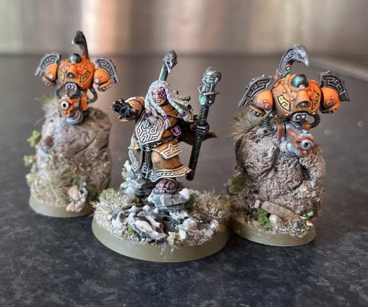 3 tabletop gaming models. Central model is a long haired dwarf in ornamented robes with outstretched hand and techno-stave, levitating on rocks. Flanked on both sides by hovering drones of roughly torso shape with guns hanging below them.