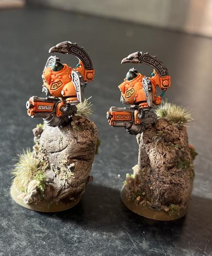 Profile view of two painted tabletop miniatures. They are floating mechanical drones in roughly the shape of a torso carrying a gun each underneath them. They are positioned over boulders