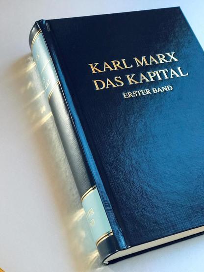 Foto von "Das Kapital", Erster Band, von Karl Marx. Blauer EInband mit goldener Prägeschrift, die sich auf dem glänzenden Tisch spiegelt.