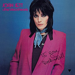Album cover for Joan Jett and the Blackhearts' "I Love Rock-n-Roll." The cover image shows Joan Jett with long black hair wearing a pinkish-purple suit coat over a black shirt with a blue scarf. She is standing against a bluish-grey wall. In the upper left hand corner, it reads "Joan Jett & the Blackhearts" with her name in all capital letters and "the Blackhearts" in all lowercase letters. In the lower right hand corner it reads "I Love Rock-n-Roll" in stylized black cursive.