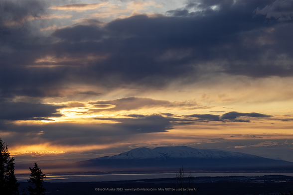 Dramatic orange sunset light to the lower left with cloud bands, light reflection the waters of Cook Inlet with Sleeping Lady on the horizon.