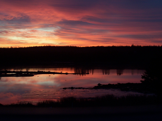 Looking over a body of water, the shore on the horizon is  black, as is the foreground. The sun is about to rise and has turned heavy clouds into grey-orange haze, and reflects off the surface of the water