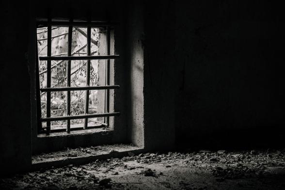 Ein vergittertes Kellerfenster durch das Licht fällt. Schwarz weiß Fotografie.