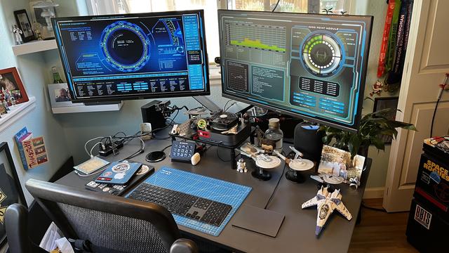 The image depicts a well-organized and tech-oriented workspace. The desk is equipped with two large monitors displaying futuristic, digital interfaces with blue and green hues, suggesting a focus on technology or gaming. The left monitor shows a circular interface with various data points, while the right monitor displays a grid-like interface with graphs and numerical data.

The desk is cluttered with various items, including a keyboard, a mouse, a calculator, and a blue cutting mat. There are also several small figurines and models, including a Star Wars spaceship and other collectibles, indicating a possible interest in science fiction or space exploration. A speaker is visible, suggesting an interest in audio or music.