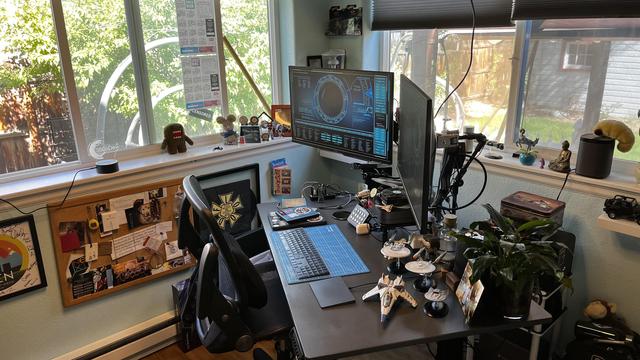 The image depicts a well-organized home office setup. The desk is positioned near a large window, allowing natural light to fill the space. On the desk, there are two computer monitors, a keyboard, and a mouse, with a blue keyboard cover. Various small figurines and models, including spacecraft, are arranged on the desk, adding a personal touch. A microphone is set up on a stand, suggesting the space is used for streaming or recording. A potted plant adds a touch of greenery to the workspace. The wall behind the desk features a bulletin board with notes and pictures, and a calendar is visible on the window sill. The overall setup is neat and functional, with a comfortable black office chair positioned in front of the desk.