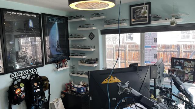 The image depicts a room with a light blue wall, featuring a workspace setup. On the left, there are two framed posters: "The Star Wars Saga Continues" and "Return of the Jedi," along with a collection of Star Wars memorabilia, including model ships and action figures, displayed on white shelves. Below the posters, there are two backpacks with patches hanging on a metal rack. The room is equipped with a large desk that holds multiple computer monitors, with one prominently displaying a yellow triangle sticker. The desk is cluttered with various items, including a camera setup and cables. A large window with a view of a wooden fence and a chain-link fence is visible, allowing natural light to enter the room. The ceiling features a circular light fixture, and a framed picture of a planet is on the wall above the shelves.