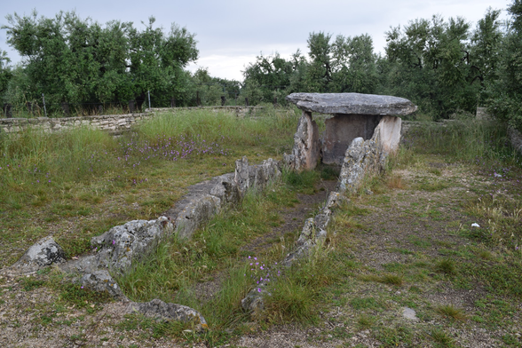 Foto do  Dolmen della Chianca, estrutura megalítica da Idade do Bronze composta por grandes pedras verticais sustentando uma laje horizontal, em um campo aberto na região de Apúlia, Itália. Céu nublado ao fundo e vegetação rasteira ao redor