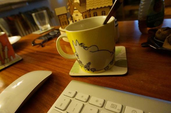 A colour photograph of a mug depicting the Moomin character Snorkfröken, or Snorkmaiden in English. The mug is on a table next to other things, a computer keyboard, a computer mouse, and in the background there is another moomin mug with hattifnattarna, or the hattifatteners in English.