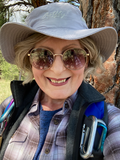 A smiling, older woman with blonde hair is in the forest wearing a beige hat, sunglasses, flannel shirt with a blue shirt beneath, a vest over both, the straps on her pack visible.