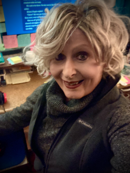 A smiling, older woman with blonde hair is turned, her desk and computer visible behind. She's wearing a charcoal, cowl-neck sweater with a vest over it.